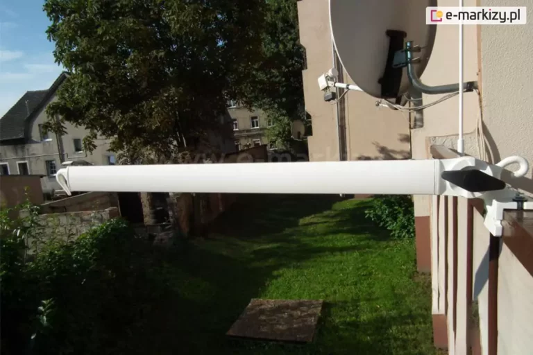 Zdjęcie prezentuje białe, aluminiowe ramię, w które wyposażona jest markiza balkonowo-poręczowa, o długości 70 cm, zamontowane prostopadle do balustrady balkonu. Ramię o prostokątnym przekroju jest wysunięte poziomo w stronę podwórza, a jego zakończenie posiada zintegrowany, wygięty hak skierowany do dołu, przeznaczony do zaczepienia dolnej rury obciążającej poszycie markizy. Konstrukcja ramienia jest zamocowana do brązowej, metalowej poręczy za pomocą białego uchwytu zaciskowego, który stabilizuje ramię dzięki czarnemu pokrętłu regulacyjnemu w kształcie rombu. W tle widoczna jest elewacja budynku w kolorze piaskowym z zamontowaną dużą, białą anteną satelitarną oraz fragmenty sąsiedniej zabudowy i gęste korony drzew. Poniżej balustrady rozciąga się zielony trawnik, na który pada cień rzucany przez konstrukcję balkonu. Po prawej stronie kadru widoczna jest ściana budynku z pionowym przewodem oraz fragment osłony balkonowej w kolorze beżowym, zamocowanej do szczebli barierki. Całość ujęcia wykonana jest przy ostrym, naturalnym świetle słonecznym, które wyraźnie eksponuje matową powierzchnię lakierowanego ramienia o długości 70 cm oraz detale techniczne systemu mocowania.