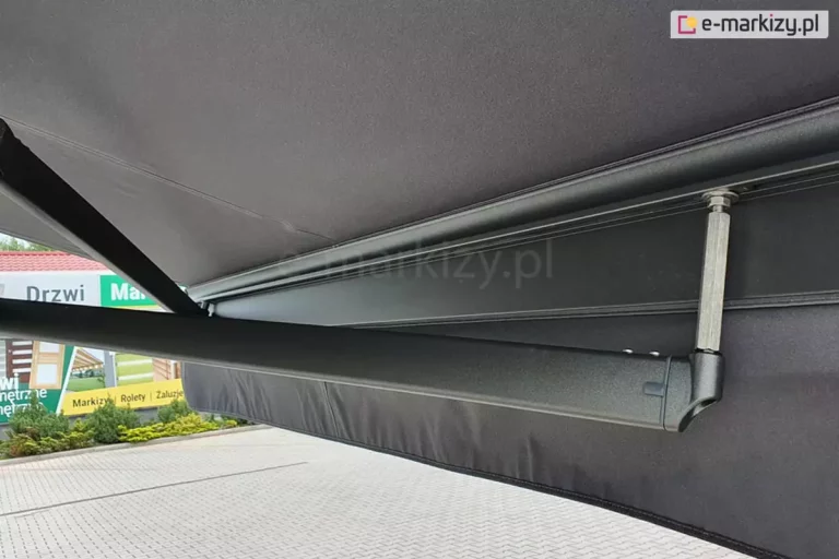Ujęta od boku, od strony obniżonego ramienia krzyżowego belka przednia markizy Palladio Aluprof widoczna od wnętrza markizy w kolorze grafitowym konstrukcji i poszycia z prostą falbaną. Wyraźnie widoczne skrzyżowanie ramion oraz wydłużone mocowanie jednego z ramion w belce przedniej.