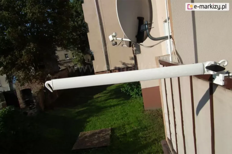Zdjęcie prezentuje ramię, w które wyposażona jest markiza balkonowo-poręczowa, ustawione w pozycji niemal poziomej, prostopadle do balustrady balkonu. Białe ramię o dlugosci 70 cm, wykonane z lakierowanego aluminium wysunięte jest daleko poza obrys barierki, a jego zakończenie posiada charakterystyczny, zintegrowany hak skierowany ku dołowi, służący do stabilnego zaczepienia dolnej rury obciążającej poszycie. Mechanizm mocujący ramię do ciemnobrązowej poręczy składa się z białej klamry zaciskowej z widoczną śrubą montażową oraz czarnego, plastikowego pokrętła w kształcie rombu, które umożliwia blokadę ramienia w wybranej pozycji. Poniżej poręczy widoczna jest jasnobeżowa osłona balkonowa zamontowana na pionowych szczeblach balustrady. W tle zdjęcia znajduje się elewacja sąsiedniego budynku w ciepłym, beżowym odcieniu z zamontowaną białą anteną satelitarną typu offset, a poniżej rozpościera się widok na zielony trawnik z cieniem rzucanym przez barierki oraz fragmenty gęstych koron drzew. Oświetlenie jest ostre i naturalne, co powoduje powstawanie wyraźnych cieni na ścianie budynku oraz na samej konstrukcji markizy, podkreślając funkcjonalność i prostotę systemu regulacji wysięgu.