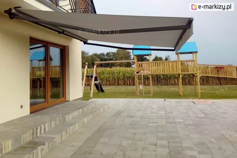 Nowoczesna markiza Australia Aluprof o wysięgu 360 cm, zamontowana na bocznej ścianie domu jednorodzinnego, osłaniająca przestronny taras wykonany z jasnoszarych płyt betonowych. Konstrukcja markizy posiada ramiona przegubowe oraz rurę nawojową w kolorze głębokiej czerni, co kontrastuje z jasną, kremową elewacją budynku. Poszycie wykonane z technicznej tkaniny w kolorze jasnoszarym jest w pełni rozwinięte, tworząc dużą strefę cienia nad wyjściem tarasowym. Markiza Australia Selt została zainstalowana nad dużymi, dwuskrzydłowymi drzwiami przesuwnymi w drewnianych ramach o ciepłym odcieniu, w których odbija się ogród. Na dalszym planie, za starannie utrzymanym trawnikiem, znajduje się rozbudowany drewniany plac zabaw dla dzieci z wieżyczkami nakrytymi niebieskimi daszkami, zjeżdżalnią oraz pomostami. Tło kompozycji stanowi wysokie pole kukurydzy oraz ściana liściastych drzew pod jasnym, lekko zachmurzonym niebem. Zdjęcie prezentuje praktyczne zastosowanie markizy tarasowej w przydomowej strefie rekreacyjnej, podkreślając jej zasięg i stabilność konstrukcji.