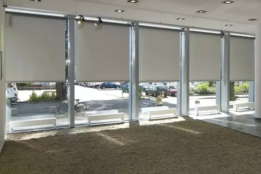 Widok z wnętrza nowoczesnego salonu wystawowego lub lobby na system wielomodułowych rolet wewnętrznych typu screen, zamontowanych w wysokich przeszkleniach fasadowych. Pięć dużych refleksoli XL z tkaniną screen w kolorze jasnoszarym zostało opuszczonych do około 3/4 wysokości okien, co zapewnia skuteczną ochronę przed słońcem przy zachowaniu naturalnego doświetlenia dolnej partii pomieszczenia. Przez przezierną strukturę tkaniny oraz odsłonięte fragmenty szyb widoczny jest parking miejski z samochodami, chodnik oraz drzewa. Wnętrze charakteryzuje się surowym designem z podłogą wysypaną drobnym żwirem lub wykładziną w kolorze brązowym oraz białymi ławkami ustawionymi wzdłuż okien. Oświetlenie sufitowe składa się z wbudowanych opraw punktowych oraz dodatkowych reflektorów kierunkowych zamontowanych na szynach przy samej konstrukcji okiennej. System sterowania roletami opiera się na mechanizmach korbowych, widocznych przy niektórych modułach po prawej stronie.