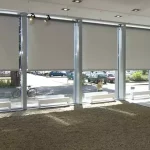 Widok z wnętrza nowoczesnego salonu wystawowego lub lobby na system wielomodułowych rolet wewnętrznych typu screen, zamontowanych w wysokich przeszkleniach fasadowych. Pięć dużych refleksoli XL z tkaniną screen w kolorze jasnoszarym zostało opuszczonych do około 3/4 wysokości okien, co zapewnia skuteczną ochronę przed słońcem przy zachowaniu naturalnego doświetlenia dolnej partii pomieszczenia. Przez przezierną strukturę tkaniny oraz odsłonięte fragmenty szyb widoczny jest parking miejski z samochodami, chodnik oraz drzewa. Wnętrze charakteryzuje się surowym designem z podłogą wysypaną drobnym żwirem lub wykładziną w kolorze brązowym oraz białymi ławkami ustawionymi wzdłuż okien. Oświetlenie sufitowe składa się z wbudowanych opraw punktowych oraz dodatkowych reflektorów kierunkowych zamontowanych na szynach przy samej konstrukcji okiennej. System sterowania roletami opiera się na mechanizmach korbowych, widocznych przy niektórych modułach po prawej stronie.