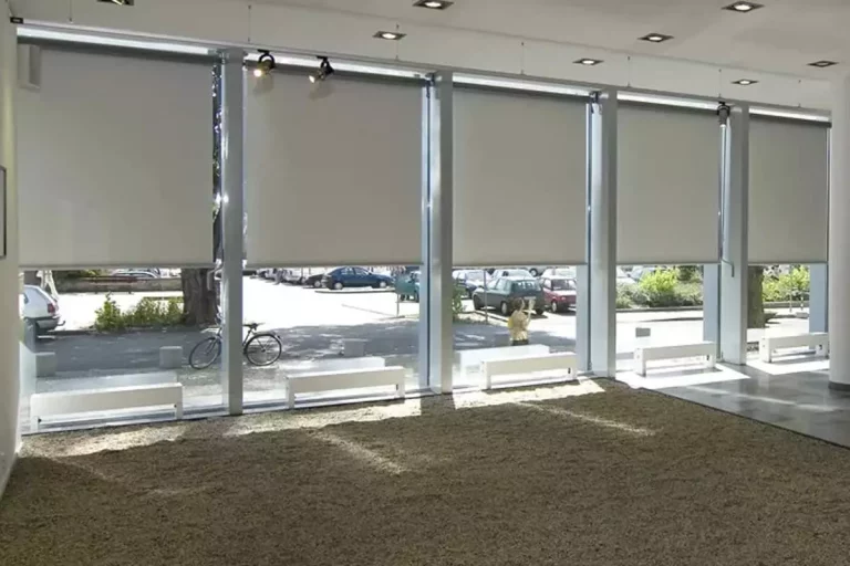Widok z wnętrza nowoczesnego salonu wystawowego lub lobby na system wielomodułowych rolet wewnętrznych typu screen, zamontowanych w wysokich przeszkleniach fasadowych. Pięć dużych refleksoli XL z tkaniną screen w kolorze jasnoszarym zostało opuszczonych do około 3/4 wysokości okien, co zapewnia skuteczną ochronę przed słońcem przy zachowaniu naturalnego doświetlenia dolnej partii pomieszczenia. Przez przezierną strukturę tkaniny oraz odsłonięte fragmenty szyb widoczny jest parking miejski z samochodami, chodnik oraz drzewa. Wnętrze charakteryzuje się surowym designem z podłogą wysypaną drobnym żwirem lub wykładziną w kolorze brązowym oraz białymi ławkami ustawionymi wzdłuż okien. Oświetlenie sufitowe składa się z wbudowanych opraw punktowych oraz dodatkowych reflektorów kierunkowych zamontowanych na szynach przy samej konstrukcji okiennej. System sterowania roletami opiera się na mechanizmach korbowych, widocznych przy niektórych modułach po prawej stronie.