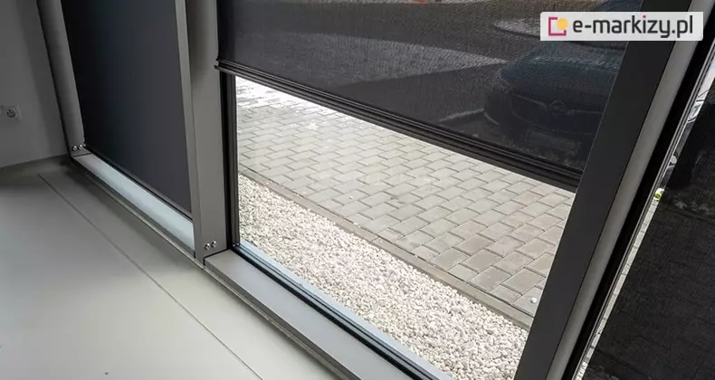 Zbliżenie wewnętrznej ramy okna w kolorze szarym, w którym zastosowany został refleksol 85A z tkanią screen. Środkowa część okna została nieco odsłonięta, dzięki czemu widoczny jest chodnik oraz przestrzeń przy ścianie budynku wysypana drobnymi jasnymi kamykami.