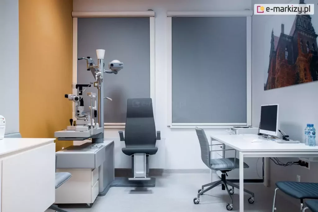 Nowoczesny gabinet medyczny wyposażony w stanowisko do diagnostyki okulistycznej z urządzeniem do badania oczu oraz ergonomicznym fotelem. Po prawej stronie znajduje się białe biurko z komputerem i krzesłem na kółkach. Na ścianie zawieszono wydruk fotograficzny przedstawiający zabytkowy budynek. Duże okna zasłonięte są wewnętrznymi roletami Refleksol 90 Selt, montowanymi w estetycznych, zaokrąglonych kasetach w kolorze białym. Tkanina o szarym odcieniu rozprasza światło dzienne, ograniczając olśnienie i poprawiając warunki pracy z komputerem i sprzętem medycznym.