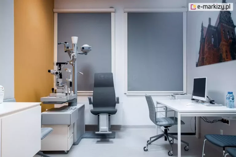 Nowoczesny gabinet medyczny wyposażony w stanowisko do diagnostyki okulistycznej z urządzeniem do badania oczu oraz ergonomicznym fotelem. Po prawej stronie znajduje się białe biurko z komputerem i krzesłem na kółkach. Na ścianie zawieszono wydruk fotograficzny przedstawiający zabytkowy budynek. Duże okna zasłonięte są wewnętrznymi roletami Refleksol 90 Selt, montowanymi w estetycznych, zaokrąglonych kasetach w kolorze białym. Tkanina o szarym odcieniu rozprasza światło dzienne, ograniczając olśnienie i poprawiając warunki pracy z komputerem i sprzętem medycznym.