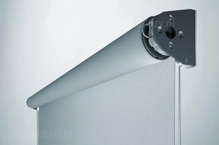 Zbliżenie na górną część refleksola XL MA Aluprof, prezentujące detale mechanizmu sterującego i mocowania. Zdjęcie przedstawia aluminiową rurę nawojową z nawiniętą jasnoszarą tkaniną o drobnoziarnistej strukturze, osadzoną w ciemnym, metalowym wsporniku bocznym. Widoczny jest plastikowy mechanizm koralikowy z przezroczystym łańcuszkiem kulkowym służącym do podnoszenia i opuszczania rolety. Tło jest jasne i jednolite, podkreślające techniczne szczegóły spasowania elementów systemu.
