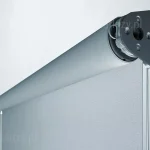 Zdjęcie prezentuje szczegółowy widok techniczny górnego mechanizmu refleksola XL Aluprof w ujęciu makro. Widoczny jest boczny, stalowy uchwyt montażowy w kolorze ciemnoszarym o charakterystycznym, trapezoidalnym kształcie, przytwierdzony za pomocą śrub do zakończenia rury nawojowej. Na wałku nawinięta jest tkanina techniczna o wyraźnym splocie siatkowym w kolorze jasnoszarym, która opada pionowo w dół, tworząc gładką płaszczyznę osłony. Detal ukazuje również mechanizm obrotowy osadzony wewnątrz rury oraz sposób wyprowadzenia krawędzi materiału. Całość kompozycji utrzymana jest w chłodnej, profesjonalnej kolorystyce na jasnym, neutralnym tle, co podkreśla nowoczesną konstrukcję i wysoką jakość wykonania komponentów systemu osłonowego.