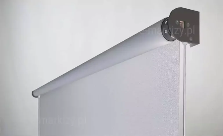 Techniczna prezentacja detali konstrukcyjnych refleksola XS Aluprof na prowadnicach linkowych. Zdjęcie ukazuje górną część systemu, gdzie srebrna rura nawojowa łączy się z ciemnym, solidnym wspornikiem bocznym. Jasnoszary materiał tekstylny jest częściowo rozwinięty, ukazując swoją strukturę przepuszczającą światło, przy jednoczesnym zachowaniu ochrony przed nagrzewaniem. Kluczowym elementem jest boczna linka napinająca, która przechodzi przez dolne obciążenie tkaniny (widoczne jako subtelne zakończenie krawędzi), co pozwala na montaż rolety w miejscach, gdzie nie ma możliwości zastosowania klasycznych prowadnic bocznych.