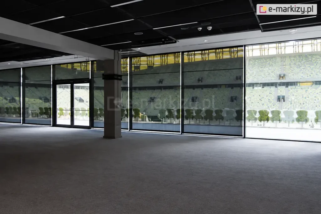 Szeroki widok z wnętrza dużej, nowoczesnej hali lub loży VIP na stadionie sportowym, gdzie na przeszklonej ścianie zainstalowano wielkoformatowe rolety przeciwsłoneczne typu screen Aluprof. Ciemne, półtransparentne tkaniny są całkowicie rozwinięte, zasłaniając wysokie okna i drzwi wyjściowe na trybuny. Przez strukturę materiału refleksoli XL wyraźnie widoczny jest widok na stadion: puste rzędy krzesełek w kolorze żółtym i zielonym oraz fragmenty konstrukcji dachu i oświetlenia obiektu. Wewnątrz pomieszczenia panuje półmrok, podkreślający skuteczność ochrony przed nadmiernym blaskiem, przy zachowaniu pełnej widoczności na zewnątrz. Sala posiada surowy, industrialny sufit z widocznymi instalacjami oraz betonowy słup konstrukcyjny. Światło dzienne wpada do wnętrza jedynie przez szczeliny między poszczególnymi modułami refleksoli, tworząc pionowe linie jasnego światła na podłodze o szarej wykładzinie.