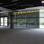 Szeroki widok z wnętrza dużej, nowoczesnej hali lub loży VIP na stadionie sportowym, gdzie na przeszklonej ścianie zainstalowano wielkoformatowe rolety przeciwsłoneczne typu screen Aluprof. Ciemne, półtransparentne tkaniny są całkowicie rozwinięte, zasłaniając wysokie okna i drzwi wyjściowe na trybuny. Przez strukturę materiału refleksoli XL wyraźnie widoczny jest widok na stadion: puste rzędy krzesełek w kolorze żółtym i zielonym oraz fragmenty konstrukcji dachu i oświetlenia obiektu. Wewnątrz pomieszczenia panuje półmrok, podkreślający skuteczność ochrony przed nadmiernym blaskiem, przy zachowaniu pełnej widoczności na zewnątrz. Sala posiada surowy, industrialny sufit z widocznymi instalacjami oraz betonowy słup konstrukcyjny. Światło dzienne wpada do wnętrza jedynie przez szczeliny między poszczególnymi modułami refleksoli, tworząc pionowe linie jasnego światła na podłodze o szarej wykładzinie.
