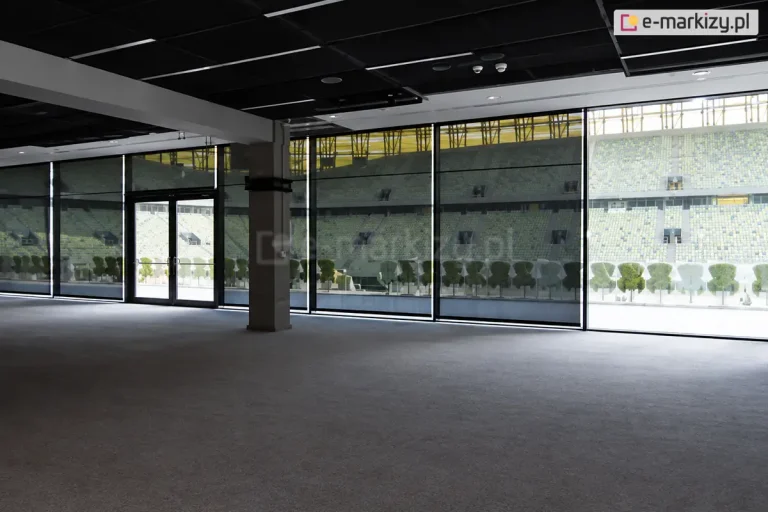 Szeroki widok z wnętrza dużej, nowoczesnej hali lub loży VIP na stadionie sportowym, gdzie na przeszklonej ścianie zainstalowano wielkoformatowe rolety przeciwsłoneczne typu screen Aluprof. Ciemne, półtransparentne tkaniny są całkowicie rozwinięte, zasłaniając wysokie okna i drzwi wyjściowe na trybuny. Przez strukturę materiału refleksoli XL wyraźnie widoczny jest widok na stadion: puste rzędy krzesełek w kolorze żółtym i zielonym oraz fragmenty konstrukcji dachu i oświetlenia obiektu. Wewnątrz pomieszczenia panuje półmrok, podkreślający skuteczność ochrony przed nadmiernym blaskiem, przy zachowaniu pełnej widoczności na zewnątrz. Sala posiada surowy, industrialny sufit z widocznymi instalacjami oraz betonowy słup konstrukcyjny. Światło dzienne wpada do wnętrza jedynie przez szczeliny między poszczególnymi modułami refleksoli, tworząc pionowe linie jasnego światła na podłodze o szarej wykładzinie.