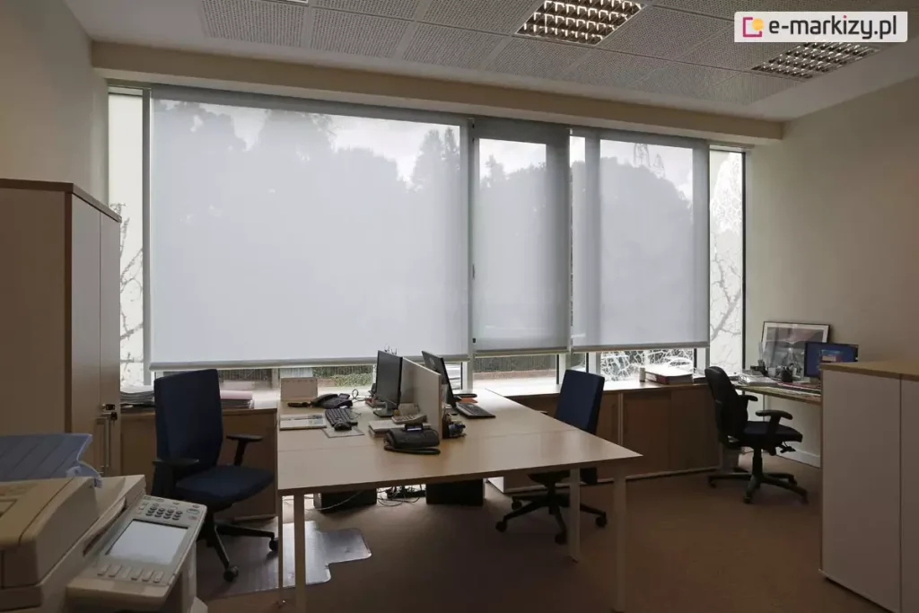 Widok z wnętrza biura na system wewnętrznych rolet przeciwsłonecznych typu screen, zamontowanych na szerokim przeszkleniu. Trzy moduły refleksola XL Aluprof z tkaniną screen w kolorze jasnoszarym, są rozwinięte na różnych wysokościach, co obrazuje możliwość indywidualnej regulacji stopnia zacienienia stanowisk pracy. Tkanina o strukturze drobnej siatki skutecznie rozprasza światło dzienne, eliminując odblaski na monitorach przy zachowaniu widoczności zarysów drzew za oknem. W pomieszczeniu znajdują się biurka z jasnym blatem, wyposażone w komputery i telefony stacjonarne, oraz ergonomiczne fotele biurowe w kolorze granatowym i czarnym. Wyposażenie wnętrza uzupełniają jasne szafy biurowe, urządzenie wielofunkcyjne na pierwszym planie oraz sufit kasetonowy z wbudowanymi oprawami oświetleniowymi typu raster.
