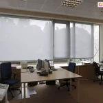 Widok z wnętrza biura na system wewnętrznych rolet przeciwsłonecznych typu screen, zamontowanych na szerokim przeszkleniu. Trzy moduły refleksola XL Aluprof z tkaniną screen w kolorze jasnoszarym, są rozwinięte na różnych wysokościach, co obrazuje możliwość indywidualnej regulacji stopnia zacienienia stanowisk pracy. Tkanina o strukturze drobnej siatki skutecznie rozprasza światło dzienne, eliminując odblaski na monitorach przy zachowaniu widoczności zarysów drzew za oknem. W pomieszczeniu znajdują się biurka z jasnym blatem, wyposażone w komputery i telefony stacjonarne, oraz ergonomiczne fotele biurowe w kolorze granatowym i czarnym. Wyposażenie wnętrza uzupełniają jasne szafy biurowe, urządzenie wielofunkcyjne na pierwszym planie oraz sufit kasetonowy z wbudowanymi oprawami oświetleniowymi typu raster.