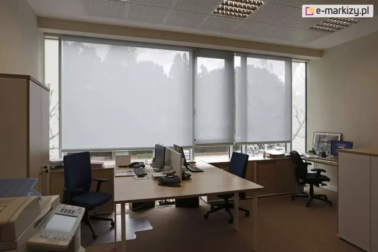 Widok z wnętrza biura na system wewnętrznych rolet przeciwsłonecznych typu screen, zamontowanych na szerokim przeszkleniu. Trzy moduły refleksola XL Aluprof z tkaniną screen w kolorze jasnoszarym, są rozwinięte na różnych wysokościach, co obrazuje możliwość indywidualnej regulacji stopnia zacienienia stanowisk pracy. Tkanina o strukturze drobnej siatki skutecznie rozprasza światło dzienne, eliminując odblaski na monitorach przy zachowaniu widoczności zarysów drzew za oknem. W pomieszczeniu znajdują się biurka z jasnym blatem, wyposażone w komputery i telefony stacjonarne, oraz ergonomiczne fotele biurowe w kolorze granatowym i czarnym. Wyposażenie wnętrza uzupełniają jasne szafy biurowe, urządzenie wielofunkcyjne na pierwszym planie oraz sufit kasetonowy z wbudowanymi oprawami oświetleniowymi typu raster.