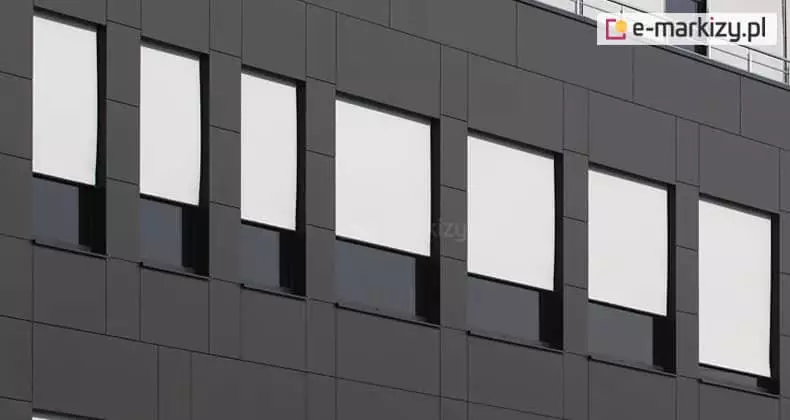 Fragment nowoczesnej elewacji budynku biurowego wyłożonej ciemnoszarymi panelami elewacyjnymi o matowym wykończeniu. W regularnych odstępach rozmieszczonych jest siedem pionowych, prostokątnych okien, w których zamontowano refleksole XS z tkaniną w kolorze jasnoszarym lub niemal białym. Przesłony są rozwinięte do około 2/3 wysokości każdego okna, tworząc rytmiczny, geometryczny wzór na fasadzie. Jasna tkanina rolet wyraźnie kontrastuje z ciemną stolarką okienną oraz antracytowym kolorem płyt elewacyjnych. W dolnych częściach okien, poniżej linii rolet, widoczne są ciemne szyby odbijające otoczenie. W prawym górnym rogu kadru widoczny jest fragment metalowej barierki zabezpieczającej dach lub taras techniczny. Zdjęcie wykonane jest z perspektywy dolnej, co podkreśla monumentalny charakter i nowoczesny design architektury budynku.