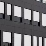 Elewacja nowoczesnego budynku z dużą liczbą prostokątnych okien, na których zamontowano zewnętrzne refleksole podtynkowe. Rolety są całkowicie rozwinięte, wykonane z jasnej tkaniny o wysokim stopniu rozproszenia światła. Kasety rolet oraz prowadnice są wkomponowane w czarne obramowania okien, co tworzy spójny i estetyczny wygląd całej fasady. Tkaniny są napięte i płasko przylegają do prowadnic, co świadczy o systemie typu ZIP, zapewniającym odporność na wiatr i dokładne zaciemnienie.