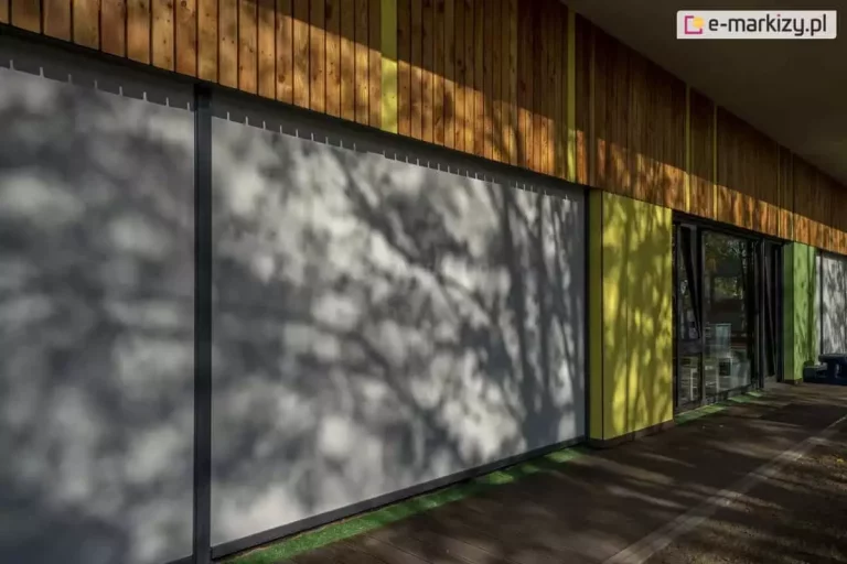 Widok zewnętrzny nowoczesnej elewacji budynku z zainstalowanym systemem wielkogabarytowych rolet zewnętrznych typu screen - refleksol Ziiip 120 Selt. Rolety w kolorze jasnoszarym są całkowicie opuszczone, zasłaniając duże przeszklenia i tworząc jednolitą płaszczyznę chroniącą przed słońcem. Na napiętej tkaninie rolet widoczne są wyraźne cienie rzucane przez pobliskie drzewa, co podkreśla matową strukturę materiału. Elewacja budynku nad roletami wykończona jest pionowymi panelami z jasnego drewna, a ściany boczne między oknami pomalowano na kontrastujące kolory: żółty oraz zielony. System osłonowy posiada widoczne, pionowe prowadnice w kolorze antracytowym, które dzielą sekcje materiału. Przed budynkiem znajduje się drewniany taras oraz pas sztucznej trawy. Zdjęcie wykonane w pełnym słońcu, prezentuje estetyczne i funkcjonalne dopasowanie osłon typu screen do współczesnej architektury obiektów użyteczności publicznej.