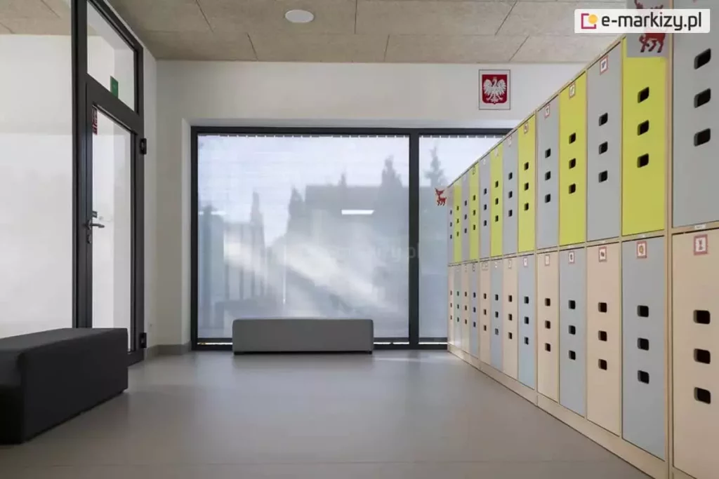 Jasne wnętrze nowoczesnej szatni szkolnej lub przedszkolnej z dużym przeszkleniem, zabezpieczonym zewnętrzną roletą materiałową typu screen. Roleta w kolorze jasnoszarym jest całkowicie rozwinięta, zasłaniając dwuskrzydłowe okno o ciemnych profilach. Dzięki właściwościom przeziernym tkaniny, mimo pełnego zasłonięcia, do wnętrza wpada rozproszone światło dzienne, a kontury budynków i drzew na zewnątrz pozostają delikatnie widoczne. Po prawej stronie kadru znajduje się ciąg pionowych szafek skrytkowych w kolorach szarym i limonkowym, ustawionych na jasnej, jednolitej podłodze. Na ścianie nad szafkami widoczny jest polski symbol narodowy (godło). Po lewej stronie znajdują się przeszklone drzwi wejściowe oraz antracytowa pufa do siedzenia. Pomieszczenie jest przestronne i minimalistyczne, a zastosowana roleta screen skutecznie chroni wnętrze przed nadmiernym nagrzewaniem i odblaskami, zachowując jednocześnie kontakt wzrokowy z otoczeniem.
