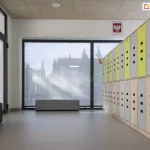 Jasne wnętrze nowoczesnej szatni szkolnej lub przedszkolnej z dużym przeszkleniem, zabezpieczonym zewnętrzną roletą materiałową typu screen. Roleta w kolorze jasnoszarym jest całkowicie rozwinięta, zasłaniając dwuskrzydłowe okno o ciemnych profilach. Dzięki właściwościom przeziernym tkaniny, mimo pełnego zasłonięcia, do wnętrza wpada rozproszone światło dzienne, a kontury budynków i drzew na zewnątrz pozostają delikatnie widoczne. Po prawej stronie kadru znajduje się ciąg pionowych szafek skrytkowych w kolorach szarym i limonkowym, ustawionych na jasnej, jednolitej podłodze. Na ścianie nad szafkami widoczny jest polski symbol narodowy (godło). Po lewej stronie znajdują się przeszklone drzwi wejściowe oraz antracytowa pufa do siedzenia. Pomieszczenie jest przestronne i minimalistyczne, a zastosowana roleta screen skutecznie chroni wnętrze przed nadmiernym nagrzewaniem i odblaskami, zachowując jednocześnie kontakt wzrokowy z otoczeniem.