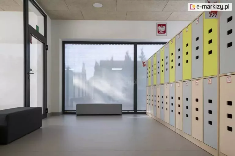 Jasne wnętrze nowoczesnej szatni szkolnej lub przedszkolnej z dużym przeszkleniem, zabezpieczonym zewnętrzną roletą materiałową typu screen. Roleta w kolorze jasnoszarym jest całkowicie rozwinięta, zasłaniając dwuskrzydłowe okno o ciemnych profilach. Dzięki właściwościom przeziernym tkaniny, mimo pełnego zasłonięcia, do wnętrza wpada rozproszone światło dzienne, a kontury budynków i drzew na zewnątrz pozostają delikatnie widoczne. Po prawej stronie kadru znajduje się ciąg pionowych szafek skrytkowych w kolorach szarym i limonkowym, ustawionych na jasnej, jednolitej podłodze. Na ścianie nad szafkami widoczny jest polski symbol narodowy (godło). Po lewej stronie znajdują się przeszklone drzwi wejściowe oraz antracytowa pufa do siedzenia. Pomieszczenie jest przestronne i minimalistyczne, a zastosowana roleta screen skutecznie chroni wnętrze przed nadmiernym nagrzewaniem i odblaskami, zachowując jednocześnie kontakt wzrokowy z otoczeniem.