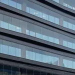 Zbliżenie na szklaną fasadę wielokondygnacyjnego budynku biurowego z widocznymi poziomymi pasami konstrukcyjnymi w kolorze czarnym lub ciemnografitowym. Za dużymi taflami szkła widoczne są wewnętrzne przesłony okienne w jasnym kolorze, które częściowo zasłaniają wnętrza poszczególnych pięter. W przeszkleniach odbija się niebo, a przez transparentne sekcje dostrzec można elementy oświetlenia sufitowego w biurach. Konstrukcja elewacji charakteryzuje się czystymi liniami i nowoczesnym, korporacyjnym designem.