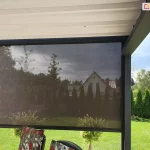 Zdjęcie prezentuje boczną osłonę przeciwsłoneczną typu refleksol zamontowaną w konstrukcji nowoczesnej pergoli tarasowej Luxury. Półtransparentna tkanina techniczna w kolorze ciemnoszarym lub antracytowym jest niemal całkowicie rozwinięta, skutecznie ograniczając nasłonecznienie przy jednoczesnym zachowaniu widoczności na zewnątrz. Materiał porusza się w pionowych prowadnicach bocznych zintegrowanych ze słupami pergoli, a jego dolna krawędź jest dociążona estetyczną listwą końcową. Powyżej widoczne jest białe, segmentowe poszycie dachowe wykonane z tkaniny, które współgra z ciemną ramą konstrukcji. W tle roztacza się widok na zadbany ogród z zielonym trawnikiem, tujami oraz fragmentem sąsiedniej zabudowy jednorodzinnej, co podkreśla funkcjonalność osłony w zapewnianiu prywatności i komfortu termicznego na tarasie.