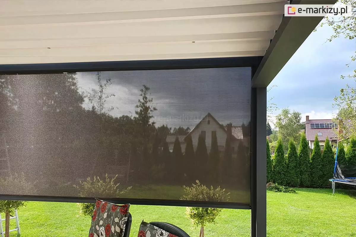 Zdjęcie prezentuje boczną osłonę przeciwsłoneczną typu refleksol zamontowaną w konstrukcji nowoczesnej pergoli tarasowej Luxury. Półtransparentna tkanina techniczna w kolorze ciemnoszarym lub antracytowym jest niemal całkowicie rozwinięta, skutecznie ograniczając nasłonecznienie przy jednoczesnym zachowaniu widoczności na zewnątrz. Materiał porusza się w pionowych prowadnicach bocznych zintegrowanych ze słupami pergoli, a jego dolna krawędź jest dociążona estetyczną listwą końcową. Powyżej widoczne jest białe, segmentowe poszycie dachowe wykonane z tkaniny, które współgra z ciemną ramą konstrukcji. W tle roztacza się widok na zadbany ogród z zielonym trawnikiem, tujami oraz fragmentem sąsiedniej zabudowy jednorodzinnej, co podkreśla funkcjonalność osłony w zapewnianiu prywatności i komfortu termicznego na tarasie.