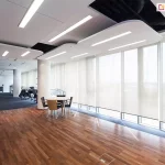 Nowoczesne wnętrze biurowe z dużą przestrzenią typu open space, podzieloną na strefy roboczą i wypoczynkową. Na pierwszym planie znajduje się niewielki stolik z czterema krzesłami ustawiony na podłodze z ciemnego drewna. W tle widoczna wykładzina biurowa w kolorze grafitowym, stanowiska pracy z monitorami i fotele biurowe. Ściana po prawej stronie to ciąg dużych okien sięgających od podłogi do sufitu, zasłoniętych roletami typu screen w kolorze jasnym, przepuszczającymi rozproszone światło dzienne. Sufit podwieszany z nowoczesnymi panelami akustycznymi i liniowym oświetleniem LED. Pomieszczenie utrzymane w minimalistycznym, profesjonalnym stylu.