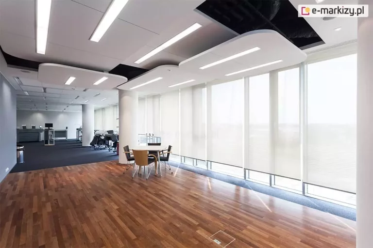 Nowoczesne wnętrze biurowe z dużą przestrzenią typu open space, podzieloną na strefy roboczą i wypoczynkową. Na pierwszym planie znajduje się niewielki stolik z czterema krzesłami ustawiony na podłodze z ciemnego drewna. W tle widoczna wykładzina biurowa w kolorze grafitowym, stanowiska pracy z monitorami i fotele biurowe. Ściana po prawej stronie to ciąg dużych okien sięgających od podłogi do sufitu, zasłoniętych roletami typu screen w kolorze jasnym, przepuszczającymi rozproszone światło dzienne. Sufit podwieszany z nowoczesnymi panelami akustycznymi i liniowym oświetleniem LED. Pomieszczenie utrzymane w minimalistycznym, profesjonalnym stylu.