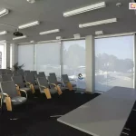 Widok na salę szkoleniową wyposażoną w system wewnętrznych rolet przeciwsłonecznych typu screen, zainstalowanych na szerokim przeszkleniu fasadowym. Cztery wielkoformatowe refleksole XL z tkaniną screen w kolorze jasnoszarym są całkowicie rozwinięte, co zapewnia jednolitą barierę wizualną i termiczną, redukując odblaski w pomieszczeniu przy jednoczesnym zachowaniu delikatnej przezierności na krajobraz zewnętrzny. Wnętrze sali obejmuje rzędy szarych foteli z drewnianymi pulpitami bocznymi, ustawionych przodem do stołu prelegenta. Na suficie widoczny jest system profesjonalnego oświetlenia liniowego oraz rzutnik multimedialny. Po bokach okien znajdują się ścianki wystawiennicze (roll-upy) z logotypami, a białe filary konstrukcyjne dzielą płaszczyznę przeszkleń na sekcje, integrując się z estetyką osłon okiennych.