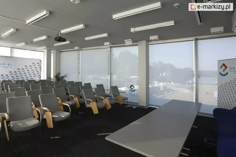 Widok na salę szkoleniową wyposażoną w system wewnętrznych rolet przeciwsłonecznych typu screen, zainstalowanych na szerokim przeszkleniu fasadowym. Cztery wielkoformatowe refleksole XL z tkaniną screen w kolorze jasnoszarym są całkowicie rozwinięte, co zapewnia jednolitą barierę wizualną i termiczną, redukując odblaski w pomieszczeniu przy jednoczesnym zachowaniu delikatnej przezierności na krajobraz zewnętrzny. Wnętrze sali obejmuje rzędy szarych foteli z drewnianymi pulpitami bocznymi, ustawionych przodem do stołu prelegenta. Na suficie widoczny jest system profesjonalnego oświetlenia liniowego oraz rzutnik multimedialny. Po bokach okien znajdują się ścianki wystawiennicze (roll-upy) z logotypami, a białe filary konstrukcyjne dzielą płaszczyznę przeszkleń na sekcje, integrując się z estetyką osłon okiennych.