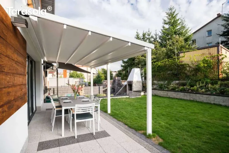 Biała pergola tarasowa Tarasola Simple o konstrukcji przyściennej zamontowana nad nowoczesnym tarasem wyłożonym jasnymi płytami chodnikowymi z ciemniejszymi pasami dekoracyjnymi. Zadaszenie pergoli wykonane jest z trójwarstwowej tkaniny technicznej ułożonej w charakterystyczne, równolegle biegnące fałdy, które są wsparte na poprzecznych profilach aluminiowych. Konstrukcja opiera się z jednej strony na elewacji budynku wykończonej panelami o strukturze drewna, a z drugiej strony na dwóch pionowych, kwadratowych słupach nośnych w kolorze białym. Pod pergolą znajduje się zestaw mebli ogrodowych składający się z dużego szarego stołu oraz sześciu białych krzeseł z ciemnymi poduszkami na siedziskach. Na stole ustawiony jest wazon z kwiatami oraz zastawa obiadowa. W tle widoczny jest zadbany trawnik, murowany grill ogrodowy o jasnej kolorystyce oraz niska roślinność i drzewa iglaste oddzielające posesję od sąsiednich budynków. Całość kompozycji przedstawiona jest w pełnym świetle dziennym przy lekko zachmurzonym niebie.