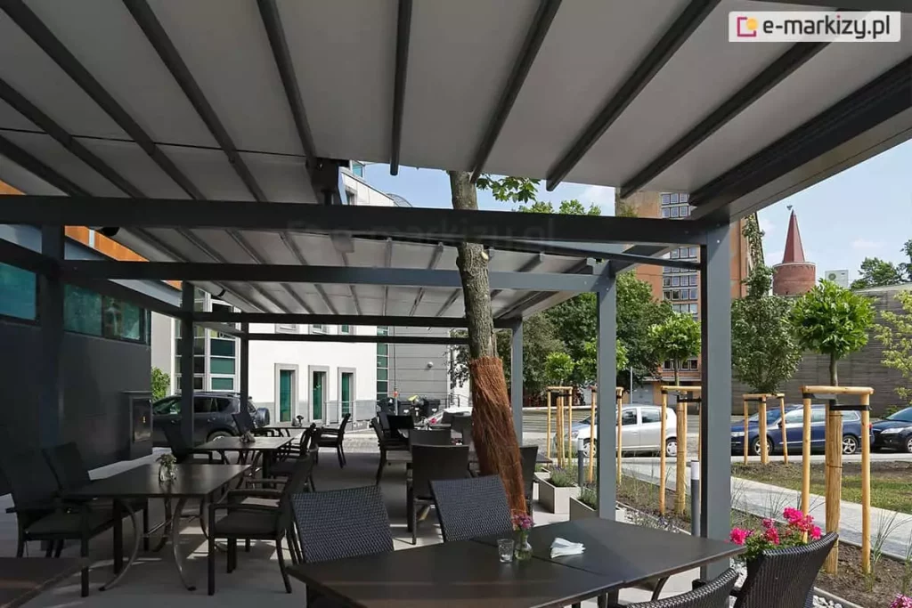 Wnętrze rozbudowanej, wielomodułowej pergoli tarasowej Solid Aluprof o konstrukcji aluminiowej w kolorze antracytowym, zaadaptowanej na ogródek restauracyjny. Zadaszenie składa się z rozsuwanego, jasnoszarego materiału zamontowanego na prowadnicach. Pod pergolą ustawiono liczne ciemne stoły z dopasowanymi fotelami z technorattanu. Konstrukcja została zaprojektowana w sposób omijający rosnące drzewo, którego pień zabezpieczono brązową matą osłonową. W tle widoczna jest nowoczesna architektura miejska: biały budynek biurowy, parking z samochodami oraz młode drzewka w ozdobnych palisadach. Po prawej stronie w oddali majaczy ceglana wieża o stożkowym dachu pod błękitnym niebem. Na stolikach restauracyjnych widoczne są drobne dekoracje, takie jak małe wazony z kwiatami.