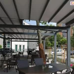 Wnętrze rozbudowanej, wielomodułowej pergoli tarasowej Solid Aluprof o konstrukcji aluminiowej w kolorze antracytowym, zaadaptowanej na ogródek restauracyjny. Zadaszenie składa się z rozsuwanego, jasnoszarego materiału zamontowanego na prowadnicach. Pod pergolą ustawiono liczne ciemne stoły z dopasowanymi fotelami z technorattanu. Konstrukcja została zaprojektowana w sposób omijający rosnące drzewo, którego pień zabezpieczono brązową matą osłonową. W tle widoczna jest nowoczesna architektura miejska: biały budynek biurowy, parking z samochodami oraz młode drzewka w ozdobnych palisadach. Po prawej stronie w oddali majaczy ceglana wieża o stożkowym dachu pod błękitnym niebem. Na stolikach restauracyjnych widoczne są drobne dekoracje, takie jak małe wazony z kwiatami.