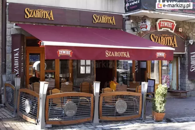 Duża tarasowa markiza Jamaica z nadrukiem reklamowym, zamontowana nad ogródkiem letnim restauracji Szarotka. Zdjęcie przedstawia w pełni rozwiniętą osłonę przeciwsłoneczną w kolorze bordowym, zamocowaną na elewacji wykonanej z szarych, kamiennych łupków. Markiza posiada prostą, długą falbanę wykończoną na dole białą lamówką. Na przedniej części falbany umieszczono napisy i logotypy: centralnie znajduje się beżowy napis „SZAROTKA”, a po bokach symetrycznie rozmieszczone logo marki „ŻYWIEC”. Nad markizą, na brązowych panelach elewacyjnych, widnieją dwa podświetlane, przestrzenne napisy z nazwą lokalu. Pod zadaszeniem zorganizowano ogródek restauracyjny z drewnianymi stolikami i krzesłami, częściowo osłonięty ozdobnymi, półokrągłymi płotkami z elementami drewna, metalowych rurek i szarych słupków. Po prawej stronie, przy wejściu do ogródka, stoi prostokątna donica z zielonym krzewem ozdobnym oraz tablica z menu lokalu. Całość ujęcia prezentuje frontową część restauracji w świetle dziennym, eksponując komercyjne zastosowanie markizy tarasowej.