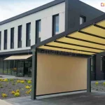 Nowoczesny budynek komercyjny o minimalistycznej architekturze, przy którym zamontowano zaawansowany system zewnętrznych osłon przeciwsłonecznych, obejmujący zarówno pergolę tarasową, jak i klasyczne markizy ścienne. Głównym elementem instalacji jest wolnostojąca pergola z wysuwanym zadaszeniem w kolorze piaskowym, wyposażona w pionowy ekran przeciwsłoneczny - zip L zip U, który zasłania część przestrzeni pod dachem, tworząc barierę przed nasłonecznieniem i wiatrem. Konstrukcja nośna pergoli wykonana została z aluminiowych profili malowanych proszkowo na ciemnoszary kolor, co doskonale koresponduje z nowoczesną bryłą budynku wykończoną w kontrastowej kolorystyce bieli i antracytu. Wzdłuż przeszklonej fasady budynku widać dodatkowe markizy kasetowe, również w kolorze piaskowym, które są zintegrowane z elewacją i zapewniają ochronę wnętrz na poziomie parteru. Budynek posiada liczne, pionowe okna o regularnym rozmieszczeniu, a przed obiektem zaprojektowano staranne otoczenie z elementami małej architektury ogrodowej, w tym wysypanymi żwirem rabatami z ozdobnymi, żółto-zielonymi krzewami. Nawierzchnia placu wykonana jest z ciemnych płyt brukowych, które harmonizują z antracytowymi akcentami elewacji oraz solidną ramą pergoli. Całość rozwiązania technicznego prezentuje wysokiej klasy systemy osłonowe, które łączą funkcjonalność ochrony przed promieniowaniem UV z nowoczesnym designem użytkowym, idealnie wpisującym się w estetykę współczesnych obiektów biurowych lub usługowych.