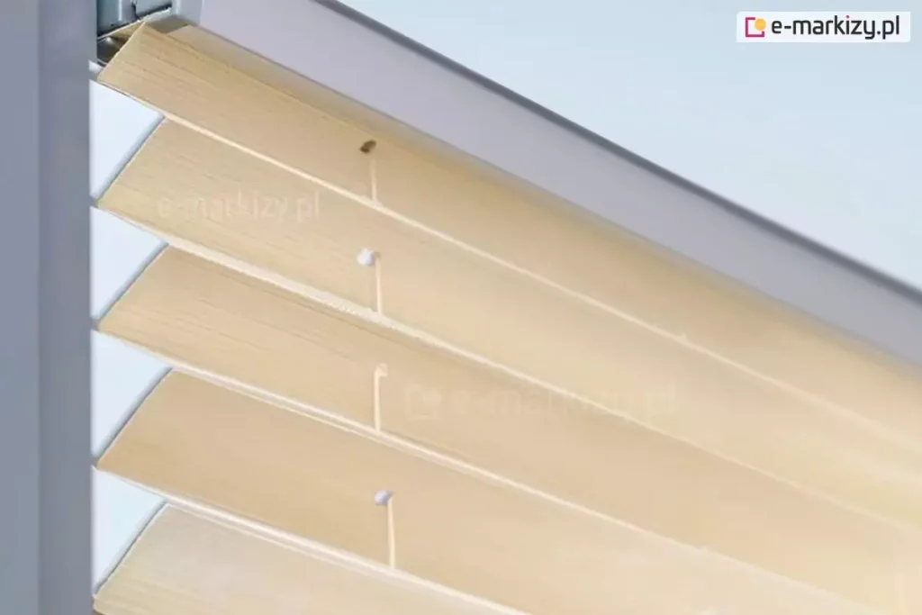 Zdjęcie prezentuje techniczne zbliżenie na górną część przesłony okiennej, którą jest żaluzja plisowana. Plisa na okno ma materiał w ciepłym, beżowym lub piaskowym odcieniu o delikatnej teksturze imitującej naturalny splot. Tkanina jest precyzyjnie zaprasowana w szerokie, regularne fałdy, które tworzą harmonijkową strukturę. W górnej części kadru widoczny jest aluminiowy profil montażowy w kolorze jasnoszarym lub srebrnym, w którym osadzona jest krawędź materiału. Przez środek zagięć tkaniny pionowo przebiega cienki, biały sznur prowadzący, przechodzący przez małe, okrągłe otwory zabezpieczone białymi wzmocnieniami. Sznurek ten jest częścią mechanizmu sterującego, który zapewnia stabilne przyleganie żaluzji do ramy okiennej i umożliwia jej płynne składanie. Ujęcie wykonane z boku, pod kątem, podkreśla trójwymiarowość załamań materiału oraz techniczne detale połączenia tkaniny z listwą górną. Całość ukazana jest na jasnym, rozmytym tle, co skupia wzrok na precyzji wykonania i estetyce osłony.