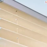 Zdjęcie prezentuje techniczne zbliżenie na górną część przesłony okiennej, którą jest żaluzja plisowana. Plisa na okno ma materiał w ciepłym, beżowym lub piaskowym odcieniu o delikatnej teksturze imitującej naturalny splot. Tkanina jest precyzyjnie zaprasowana w szerokie, regularne fałdy, które tworzą harmonijkową strukturę. W górnej części kadru widoczny jest aluminiowy profil montażowy w kolorze jasnoszarym lub srebrnym, w którym osadzona jest krawędź materiału. Przez środek zagięć tkaniny pionowo przebiega cienki, biały sznur prowadzący, przechodzący przez małe, okrągłe otwory zabezpieczone białymi wzmocnieniami. Sznurek ten jest częścią mechanizmu sterującego, który zapewnia stabilne przyleganie żaluzji do ramy okiennej i umożliwia jej płynne składanie. Ujęcie wykonane z boku, pod kątem, podkreśla trójwymiarowość załamań materiału oraz techniczne detale połączenia tkaniny z listwą górną. Całość ukazana jest na jasnym, rozmytym tle, co skupia wzrok na precyzji wykonania i estetyce osłony.