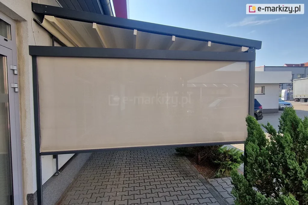 Zdjęcie przedstawia nowoczesną pergolę tkaninową w wersji przyściennej, zamontowaną na betonowym podłożu wyłożonym szarą kostką brukową. Konstrukcja nośna, w tym prowadnice dachowe oraz pionowe słupy, wykonana jest z profili aluminiowych o kwadratowym przekroju w kolorze ciemnego antracytu. Zadaszenie stanowi jasnokremowa tkanina rozpięta na poprzecznych profilach, która w tej pozycji jest całkowicie rozwinięta i tworzy lekki skos odprowadzający wodę. Na bocznej ścianie pergoli zainstalowano refleksol ZIP z tkaniną typu screen w kolorze beżowym, która jest niemal całkowicie opuszczona, zapewniając ochronę przed słońcem i wiatrem. Przez półtransparentną tkaninę rolety prześwituje zarys otoczenia, w tym zaparkowane w oddali samochody. Po lewej stronie widoczny jest fragment elewacji budynku z przeszklonymi drzwiami w szarych ramach. Po prawej stronie, wzdłuż krawędzi tarasu, rosną gęste, ciemnozielone krzewy iglaste. Całość ujęcia wykonano w pełnym świetle dziennym przy bezchmurnym, błękitnym niebie, co podkreśla funkcjonalność i estetykę osłony tarasowej w przestrzeni komercyjnej lub prywatnej.