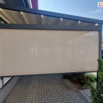 Zdjęcie przedstawia nowoczesną pergolę tkaninową w wersji przyściennej, zamontowaną na betonowym podłożu wyłożonym szarą kostką brukową. Konstrukcja nośna, w tym prowadnice dachowe oraz pionowe słupy, wykonana jest z profili aluminiowych o kwadratowym przekroju w kolorze ciemnego antracytu. Zadaszenie stanowi jasnokremowa tkanina rozpięta na poprzecznych profilach, która w tej pozycji jest całkowicie rozwinięta i tworzy lekki skos odprowadzający wodę. Na bocznej ścianie pergoli zainstalowano refleksol ZIP z tkaniną typu screen w kolorze beżowym, która jest niemal całkowicie opuszczona, zapewniając ochronę przed słońcem i wiatrem. Przez półtransparentną tkaninę rolety prześwituje zarys otoczenia, w tym zaparkowane w oddali samochody. Po lewej stronie widoczny jest fragment elewacji budynku z przeszklonymi drzwiami w szarych ramach. Po prawej stronie, wzdłuż krawędzi tarasu, rosną gęste, ciemnozielone krzewy iglaste. Całość ujęcia wykonano w pełnym świetle dziennym przy bezchmurnym, błękitnym niebie, co podkreśla funkcjonalność i estetykę osłony tarasowej w przestrzeni komercyjnej lub prywatnej.