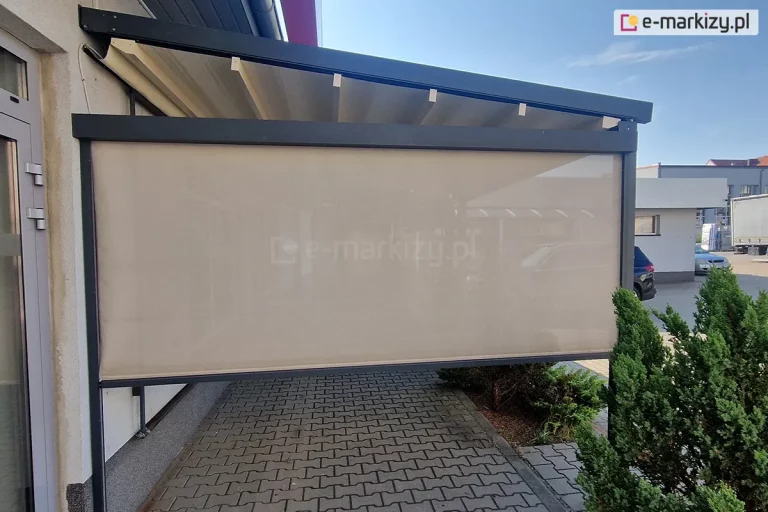 Zdjęcie przedstawia nowoczesną pergolę tkaninową w wersji przyściennej, zamontowaną na betonowym podłożu wyłożonym szarą kostką brukową. Konstrukcja nośna, w tym prowadnice dachowe oraz pionowe słupy, wykonana jest z profili aluminiowych o kwadratowym przekroju w kolorze ciemnego antracytu. Zadaszenie stanowi jasnokremowa tkanina rozpięta na poprzecznych profilach, która w tej pozycji jest całkowicie rozwinięta i tworzy lekki skos odprowadzający wodę. Na bocznej ścianie pergoli zainstalowano refleksol ZIP z tkaniną typu screen w kolorze beżowym, która jest niemal całkowicie opuszczona, zapewniając ochronę przed słońcem i wiatrem. Przez półtransparentną tkaninę rolety prześwituje zarys otoczenia, w tym zaparkowane w oddali samochody. Po lewej stronie widoczny jest fragment elewacji budynku z przeszklonymi drzwiami w szarych ramach. Po prawej stronie, wzdłuż krawędzi tarasu, rosną gęste, ciemnozielone krzewy iglaste. Całość ujęcia wykonano w pełnym świetle dziennym przy bezchmurnym, błękitnym niebie, co podkreśla funkcjonalność i estetykę osłony tarasowej w przestrzeni komercyjnej lub prywatnej.