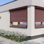 Narożnik budynku z cokołem z cegieł nad oknami, jasną elewacją oraz dwoma oknami umieszczonymi w pobliżu narożnika i drzwiami wejściowymi na lewej ścianie. Na oknach o brązowych ramach zainstalowano rolety zewnętrzne w prostokątnych skrzynkach w kolorystyce dopasowanej do barwy okien. Przed wejściem trawnik z ozdobnymi trawami przy budynku i drobnymi kamyczkami wzdłuż drogi oraz okrągłą ozdobną białą lampą. Wzdłuż prawej ściany podwórko wyłożone kostką brukową w szarym kolorze.