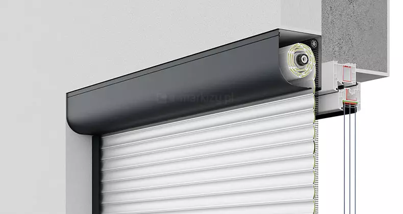 Zdjęcie przedstawia przekrój rolety zewnętrznej natynkowej w skrzyni ćwierćokrągłej. Widoczny jest zwijany pancerz aluminiowy osadzony na wale nawojowym wewnątrz kasety. Roleta może być wyposażona w napęd ręczny lub elektryczny, w zależności od potrzeb użytkownika. System montowany jest na elewacji budynku i zapewnia skuteczną ochronę przeciwsłoneczną, termiczną i antywłamaniową.