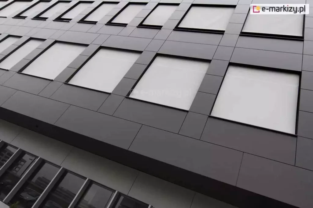 Widok z dołu na nowoczesną fasadę biurowca wyłożoną ciemnoszarymi panelami elewacyjnymi o geometrycznym układzie. W kwadratowych wnękach okiennych zainstalowano refleksole XS z tkaniną w kolorze białym lub bardzo jasnoszarym. Rolety są całkowicie opuszczone, tworząc jednolitą płaszczyznę zintegrowaną z architekturą budynku. Materiał osłon odbija światło, zapewniając ochronę termiczną wnętrz. Na niższej kondygnacji widoczne są tradycyjne przeszklenia bez osłon, w których odbija się sąsiednia zabudowa miejska. Perspektywa zdjęcia podkreśla rytmiczny układ okien i nowoczesny, industrialny charakter obiektu.