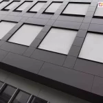 Widok z dołu na nowoczesną fasadę biurowca wyłożoną ciemnoszarymi panelami elewacyjnymi o geometrycznym układzie. W kwadratowych wnękach okiennych zainstalowano refleksole XS z tkaniną w kolorze białym lub bardzo jasnoszarym. Rolety są całkowicie opuszczone, tworząc jednolitą płaszczyznę zintegrowaną z architekturą budynku. Materiał osłon odbija światło, zapewniając ochronę termiczną wnętrz. Na niższej kondygnacji widoczne są tradycyjne przeszklenia bez osłon, w których odbija się sąsiednia zabudowa miejska. Perspektywa zdjęcia podkreśla rytmiczny układ okien i nowoczesny, industrialny charakter obiektu.