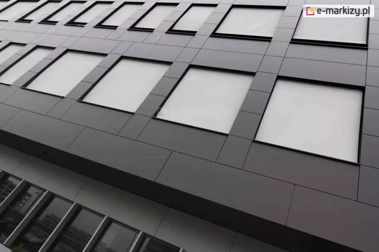 Widok z dołu na nowoczesną fasadę biurowca wyłożoną ciemnoszarymi panelami elewacyjnymi o geometrycznym układzie. W kwadratowych wnękach okiennych zainstalowano refleksole XS z tkaniną w kolorze białym lub bardzo jasnoszarym. Rolety są całkowicie opuszczone, tworząc jednolitą płaszczyznę zintegrowaną z architekturą budynku. Materiał osłon odbija światło, zapewniając ochronę termiczną wnętrz. Na niższej kondygnacji widoczne są tradycyjne przeszklenia bez osłon, w których odbija się sąsiednia zabudowa miejska. Perspektywa zdjęcia podkreśla rytmiczny układ okien i nowoczesny, industrialny charakter obiektu.