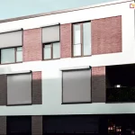Dwupiętrowy budynek mieszkalny wielorodzinny z oknami osłoniętymi roletami ZIP L. Okna pierwszego pięta przesłonięte zostały całkowicie, dwa okna na drugim piętrze po lewej zasłonięte są do połowy, a trzecie okno - po prawej stronie nie posiada zamocowanej rolety screen.