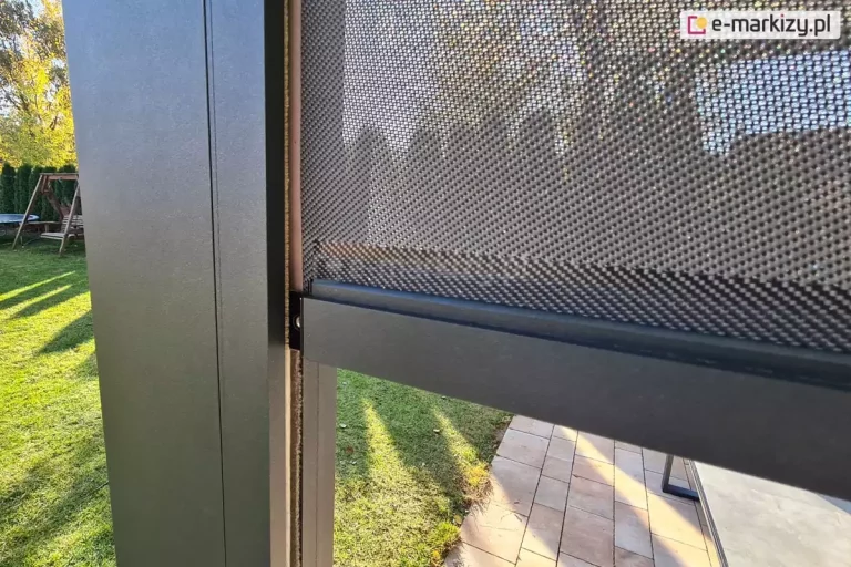 Zbliżenie na boczny profil prowadnicy oraz dolną listwę obciążającą refleksola Zip do pergoli, zamontowanego do nowoczesnej konstrukcji pergoli tarasowej. Na zdjęciu widoczna jest pionowa noga pergoli o kwadratowym przekroju, wykonana z aluminium w kolorze matowego antracytu. przy profilu słupa osadzona jest prowadnica boczna, w której porusza się tkanina techniczna o drobnym, siatkowym splocie w odcieniach szarości. Materiał rolety jest częściowo rozwinięty, a jego dolna krawędź została wykończona poziomą, masywną listwą obciążającą, dopasowaną kolorystycznie do reszty konstrukcji. Przez półtransparentną strukturę tkaniny screen oraz obok słupa prześwituje nasłoneczniony ogród z zielonym trawnikiem, fragmentem drewnianej huśtawki oraz ułożoną na tarasie jasną kostką brukową. Ostre światło słoneczne padające z boku podkreśla teksturę materiału oraz precyzyjne spasowanie elementów aluminiowych systemu osłonowego.