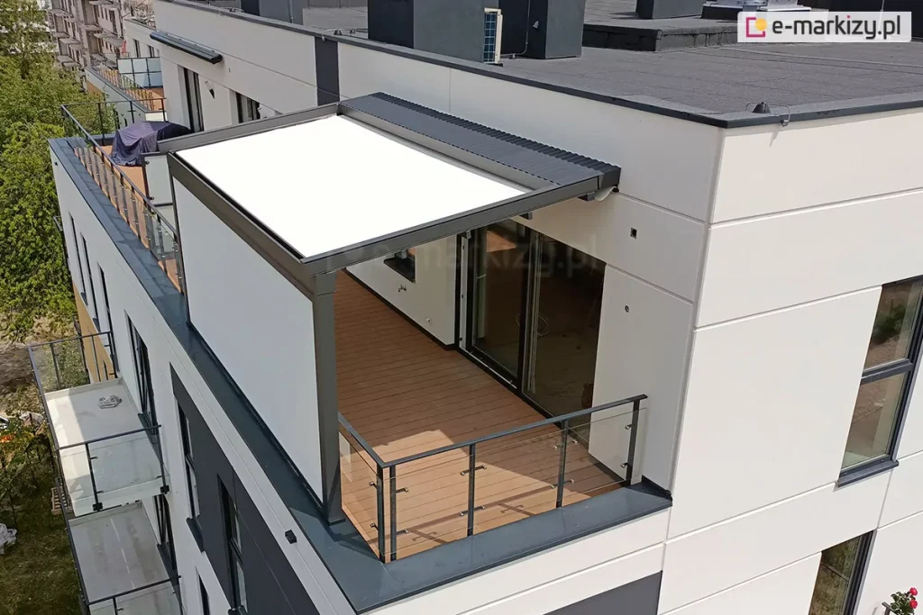 Widok z lotu ptaka na nowoczesną pergolę tarasową zainstalowaną na balkonie ostatniego piętra wielorodzinnego budynku mieszkalnego. Konstrukcja pergoli w kolorze ciemnego antracytu posiada białe, napięte zadaszenie tkaninowe o prostokątnym kształcie, które chroni część tarasu przed słońcem. Nad częścią zadaszenia znajduje się dodatkowy daszek systemowy wykonany z blachy trapezowej w kolorze konstrukcji, zabezpieczający materiał po zwinięciu. Jedna z bocznych ścian pergoli została w całości wypełniona jasną, pionową przesłoną, tworzącą osłonę od wiatru i sąsiadów. Taras wyłożony jest deskami kompozytowymi w kolorze miodowego drewna i zabezpieczony nowoczesną balustradą z metalowych profili oraz przezroczystych paneli szklanych. Na taras prowadzą duże, przeszkolone drzwi przesuwne z ciemnymi ramami. Elewacja budynku jest biała z ciemnoszarymi wstawkami między oknami, a dach budynku jest płaski, pokryty ciemną papą termozgrzewalną z widocznymi kominami i urządzeniami wentylacyjnymi. W tle widoczna jest dalsza część osiedla, balkony sąsiednich mieszkań oraz korony drzew.